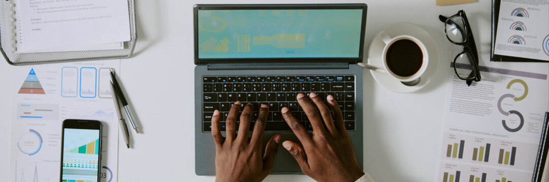 Overhead view of hands working on laptop, analyzing financial documents and graphs, with coffee, smartphone, and glasses neatly arranged on desk
