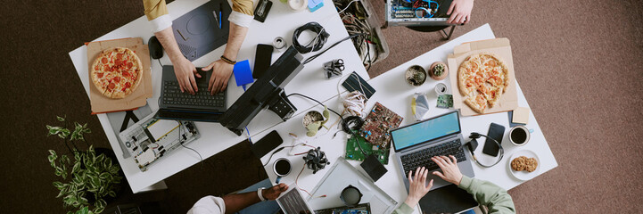 People collaborating on technology projects at office workspace, surrounding by laptops, pizza boxes, electronic components, and coffee cups. Hands visible, working on various tasks