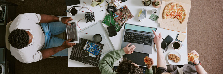 Group of people engaged in engineering projects, using various technology devices and components on table while enjoying pizza and drinks. Top-down view showing teamwork