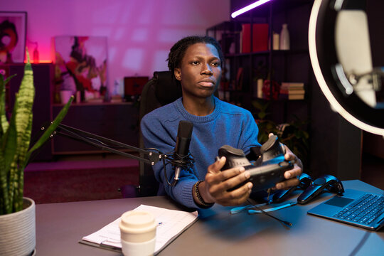 Portrait of African American man sitting at desk while holding VR headset and looking at camera while recording product review for online vlog