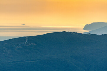 Soft golden sunset hues envelop cliffs and sea, creating calm atmosphere. Elevated perspective reveals remote winding path over lush mountain. Distant ship visible on horizon