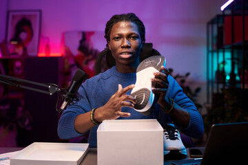 Portrait of young Black man showing fitness shoe with professionalism in modern studio setting. Background includes artwork, microphone, and shelves for an engaging atmosphere