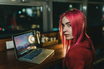 A girl with red hair and a red dress is sitting at a laptop in a cafe