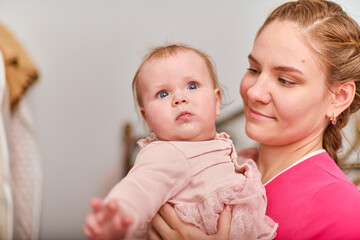 Young woman with brown hair holds blond baby girl in arms. Both appear joyful. Indoor setting with warm lighting and soft background, conveying warmth