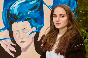 Obraz premium Young woman with long brown hair and fair skin stands beside art piece. Blue dominates painting. Outdoor setting, natural lighting enhances vivid hues