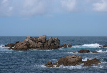 View from the Galician coast towards the Atlantic Ocean of the waves breaking against a scattered structure of rocks and foam and the blue sky with clouds separated from the water by the horizon