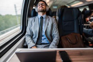 Young businessman wearing suit and tie is sitting on a train next to the window, listening to music with headphones and enjoying his comfortable trip