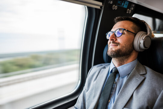 Young businessman wearing suit and tie is sitting on a train next to the window, listening to music with headphones and enjoying his comfortable trip - Powered by Adobe