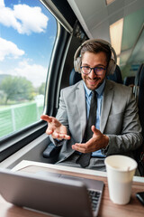 Young businessman wearing headphones and suit is making a video conference on his laptop while enjoying a cup of coffee during his train journey