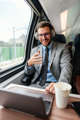 Young businessman wearing headphones and suit is making a video conference on his laptop while enjoying a cup of coffee during his train journey