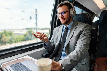 Young businessman wearing headphones and suit is making a video conference on his laptop while enjoying a cup of coffee during his train journey