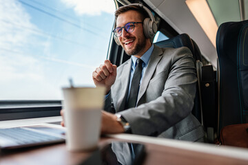 Young businessman wearing headphones and suit is making a video conference on his laptop while enjoying a cup of coffee during his train journey