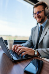 Young businessman wearing headphones and suit is making a video conference on his laptop while enjoying a cup of coffee during his train journey