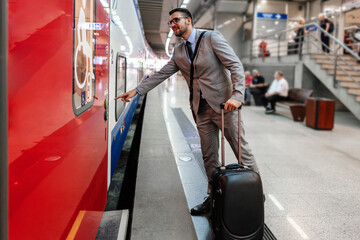 Elegant businessman waiting for a train at a bustling railway station, holding a mobile phone while pulling a trolley suitcase and gazing thoughtfully into the distance