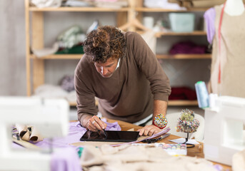 Adult man dressmaker draws pattern on tablet in sewing workshop
