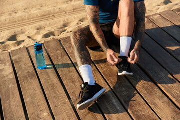 Handsome young man tying shoes while sitting on wooden deck next to sandy beach under warm sun