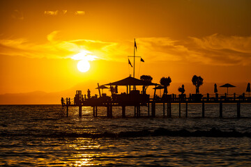 Fototapeta premium Silhouetted pier with sun loungers, trees and person watching sunset over sea and distant mountains. Side, Turkey, Mediterranean region.