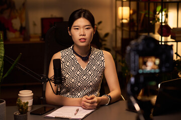 Portrait of Asian woman sitting at desk with microphone and notes conducting online video webinar with focused expression