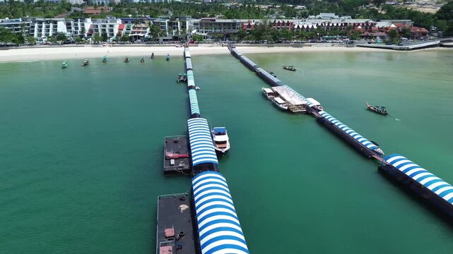 Drone flies from right to left in a half-circle around the Patong harbour, keeping the docks and pier centered while revealing the boats and scenic coastal surroundings.