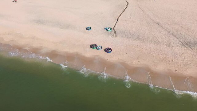Drone flies left to right in a half-circle above Mui Ne beach, filming colorful swimming buckets parked on white sand, revealing the shore, beach, and sparkling turquoise sea.