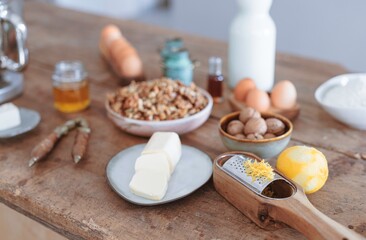 A rustic kitchen table holds an array of ingredients for baking, including vibrant lemon zest, creamy butter, and assorted nuts. The warm atmosphere invites culinary creativity