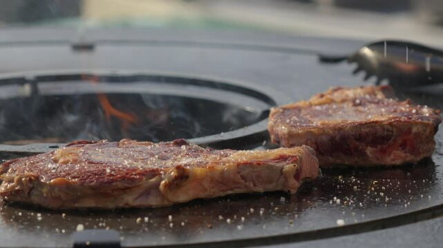 Slow motion shot of a chef adding rock salt to season steaks cooking on a bqq