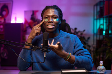 Portrait of Black man applying skincare product on face while sitting in studio setting illuminated by colorful lighting. Man is speaking into a professional microphone