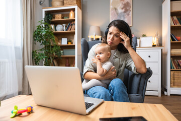 Single mother with baby on lap speaks emotionally via laptop video call to ex-husband about overdue child support, alimony, sitting in armchair wearing wireless headphones, facing financial stress.