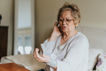 An older woman sitting in her bedroom checking her blood pressure with a digital monitor, conveying...