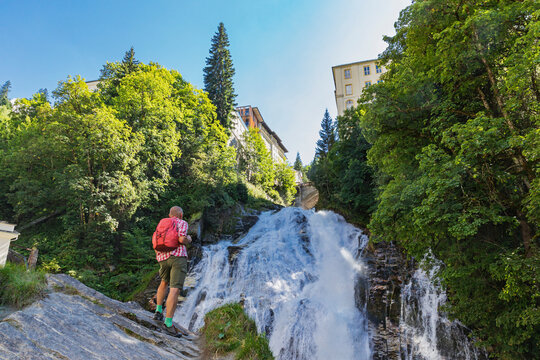 A person hiking near the Gastein Waterfall in Bad Gastein, Austria, surrounded by lush greenery and a health resort setting.
