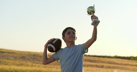 child holds rugby ball pride while raising golden trophy in sunset field. child celebrates sports achievement with trophy in hand and football under arm. child radiates joy ovictory holding trophy. - Powered by Adobe