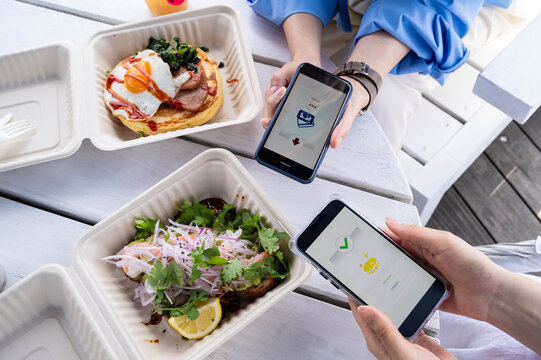 Two women complete a mobile payment transaction at an outdoor seaside café, each holding a smartphone displaying fintech apps.