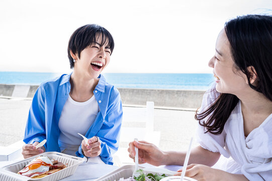 Two Japanese women smiling and laughing while sharing lunch by the ocean, symbolising strong friendship, emotional wellbeing, and healthy lifestyle