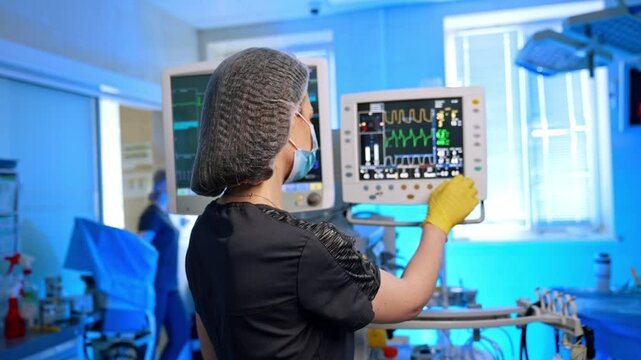 Female medic in cap and mask stands at the medical equipment switching it on. Scrub nurse turns to camera folding gloved hands on the chest.