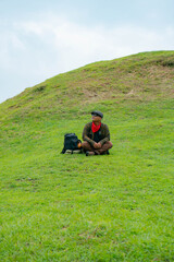An adventurer is resting by sitting on a stretch of green grass. The Asian man is enjoying the tranquility of nature by resting on a green hill that turns out to be a buried Buddhist temple.