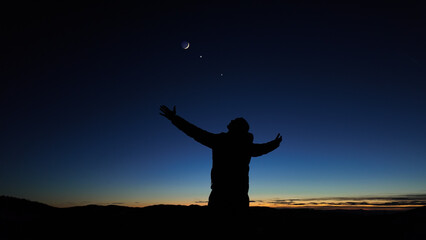 Silhouette of a man and countryside under the Moon, stars and planets in blue hour time.
