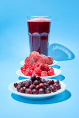 Glass with berry lemonade made from strawberries, frozen raspberries and blackcurrants. Frozen berries lie on small white plates.