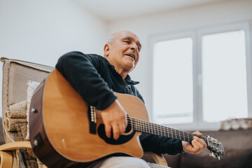 An elderly man enthusiastically plays the guitar while sitting indoors, enjoying a moment of musical creativity. The warm and inviting atmosphere complements his engaged and relaxed demeanor.