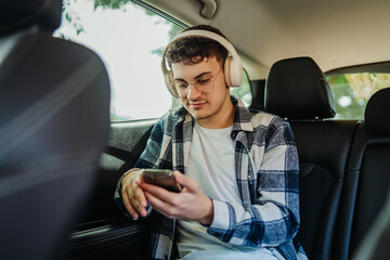 Young man listen to music on headphones and use smartphone on the backseat of the car
