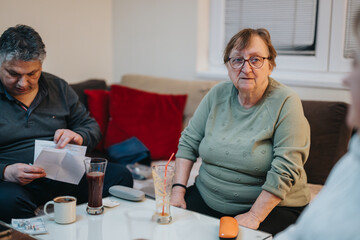 Fototapeta premium A senior woman wearing glasses is seated at a table in a cozy living room. Another individual is seen reading documents. The scene captures a homely and calm atmosphere.
