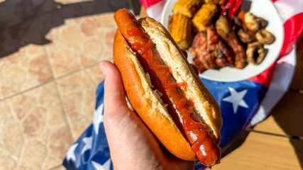 Hand grasping a ketchup-drizzled hot dog amid a sunlit American barbecue, evoking Independence Day...