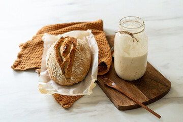 Crispy sourdough bread sits beside a glass jar filled with active starter, accompanied by a rustic cloth and a wooden board in a warm kitchen atmosphere.