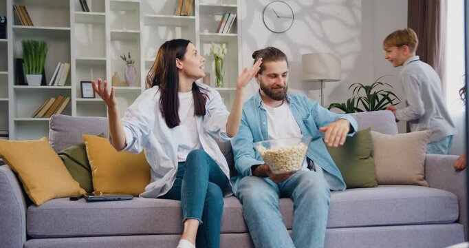 Family Father and mother sit on couch feels annoyed exhausted two joyful caucasian children running in circles and rounds in room. Upbringing difficulties, misbehaved offspring concept.