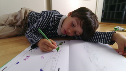 Young boy deeply engaged in drawing with a green pen while lying on the floor, focused on coloring in a book, intense creative concentration, indoor art time