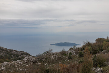 Obraz premium Distant island floats on calm sea, viewed from stony mountain ridge. Autumn foliage and rugged terrain frame the serene ocean horizon and dramatic cloud cover.