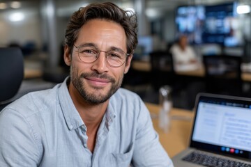 Obraz premium A young male professional smiles at the camera while working at his desk with a laptop, reflecting confidence and a positive atmosphere in a modern workspace.