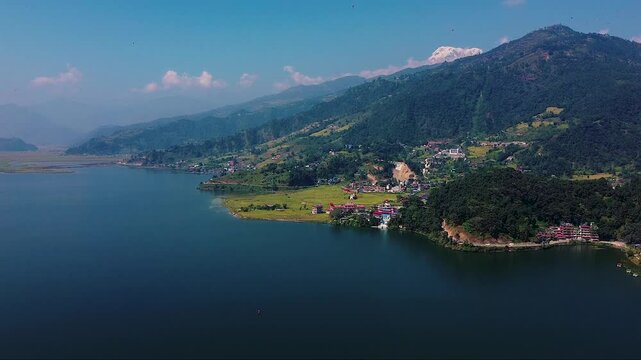 Stunning aerial view of a serene lake surrounded by lush green hills and a quaint village with colorful buildings, under a clear blue sky with scattered clouds. 