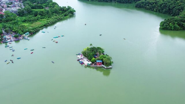Aerial view of a small island in a lake, surrounded by boats and lush greenery, with a pathway and buildings on the shore. 