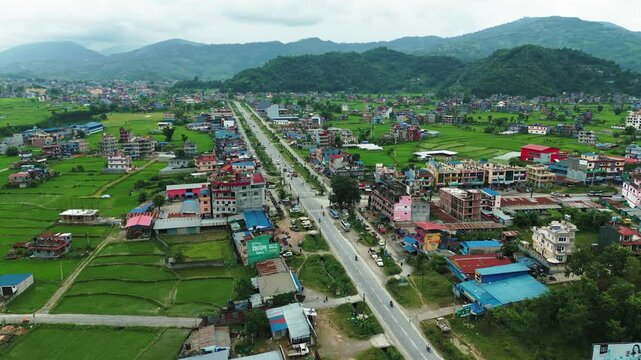 Scenic aerial shot of a peaceful village with colorful houses along a main road, surrounded by verdant fields and rugged hills.