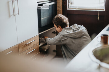 A young man wearing a casual hoodie is seen adjusting items inside a kitchen cabinet, promoting organization and a cozy sense of home.